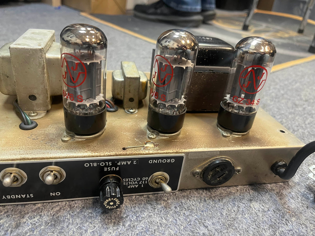 Close-up of the back panel of a 1965 Fender Deluxe Reverb AB763 (Pre-CBS Blackface, Serial A 06439) showing three vacuum tubes, power transformer, knobs, switches, and labeled controls on a gray carpeted surface.