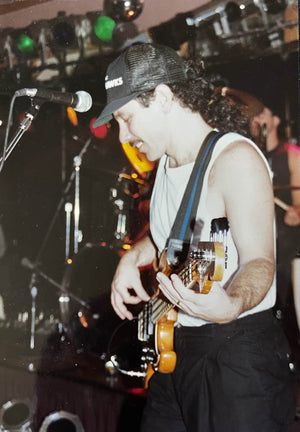 A musician in a cap and sleeveless shirt plays a Fender 1966 Jazz Bass Natural Refinish w/Case on stage, smiling, with drums and stage lights in the background.