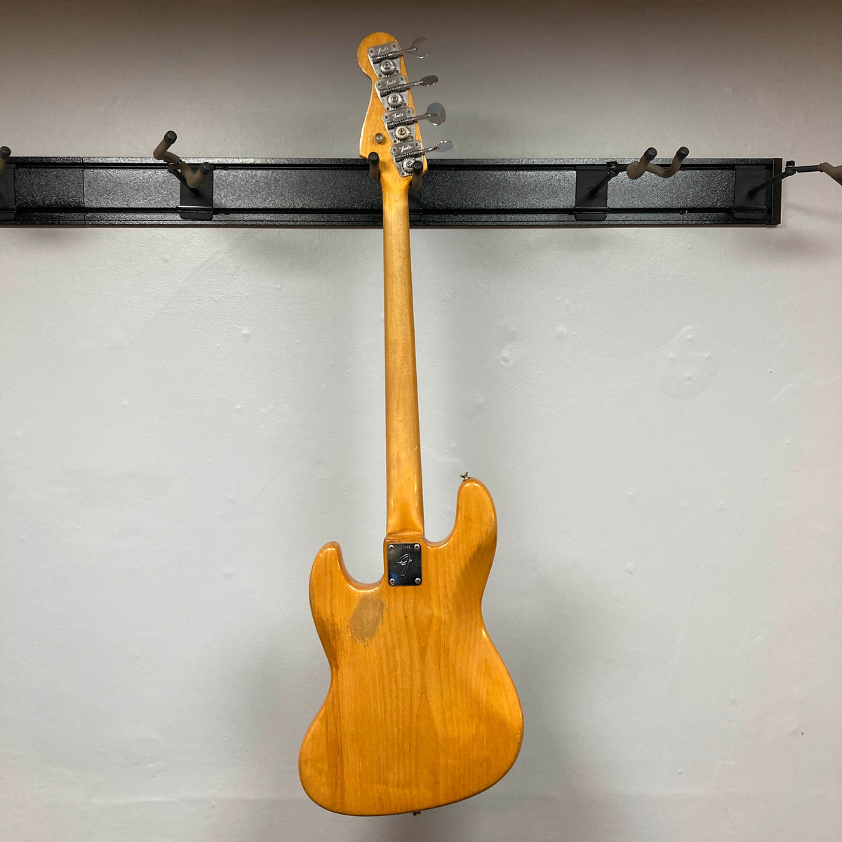 A 1966 Fender Jazz Bass Natural Refinish by Fender hangs by its headstock on a black wall rack, displayed from the back to highlight its natural finish. Includes case.