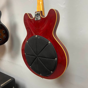 A 1967-68 Vox Sidewinder IV Bass (V272) in cherry finish hangs on the wall, showcasing a large black circular pad with visible screws on the back—classic of vintage Italian Vox bass designs.