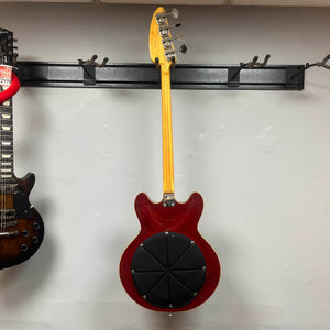 A cherry 1967-68 Vox Sidewinder IV Bass (V272) by Vox, with a circular black practice pad attached, hangs on a wall-mounted rack next to a dark electric guitar.