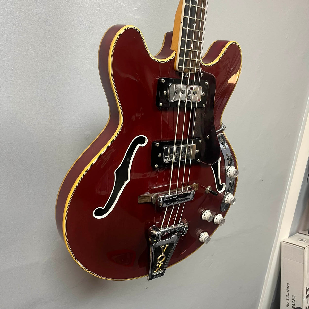 A 1967-68 Vox Sidewinder IV Bass (V272) in cherry by Vox, a semi-hollow electric bass with white trim, two humbuckers, four control knobs, and f-shaped sound holes—vintage Italian style—hangs on the wall.