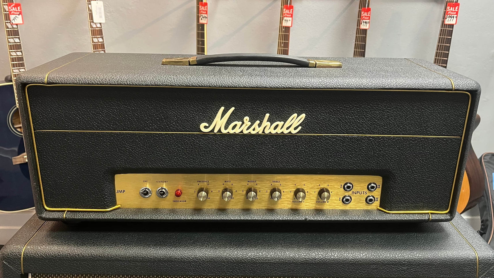 A Marshall 1971 50W Lead Head from the JMP era, complete with flight case, rests atop its cab, delivering classic British tones, with both acoustic and electric guitars hanging on the wall behind. A Marshall 1971 50W Lead Head from the JMP era, complete with flight case, rests atop its cab, delivering classic British tones, with both acoustic and electric guitars hanging on the wall behind.
