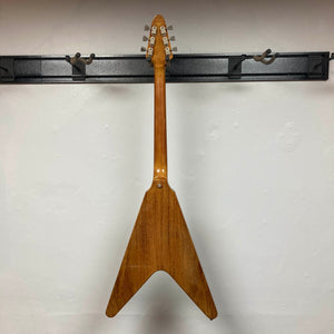 A 1975 Gibson Flying V w/Case by Gibson, featuring a natural finish, hangs by its headstock on a black wall-mounted rack, displayed from the back against a white wall.