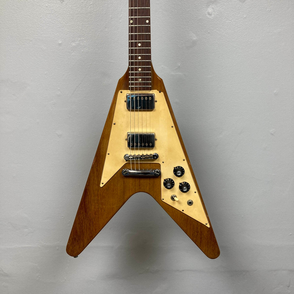 A brown 1975 Gibson Flying V electric guitar by Gibson, featuring a natural finish, distinctive V-shaped body, three control knobs, and two pickups, is displayed against a plain gray wall. Modeled after the classic vintage Flying V design, this instrument embodies the iconic style of its era and comes with a case.