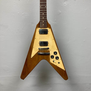 A brown 1975 Gibson Flying V electric guitar by Gibson, featuring a natural finish, distinctive V-shaped body, three control knobs, and two pickups, is displayed against a plain gray wall. Modeled after the classic vintage Flying V design, this instrument embodies the iconic style of its era and comes with a case.