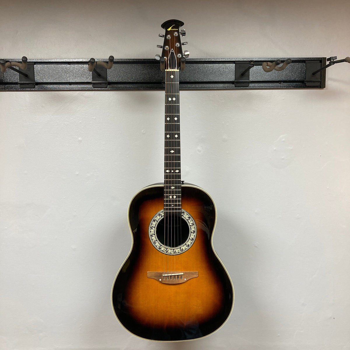 A 1976 Ovation 1112-1 Acoustic Guitar w/Case, made in the USA by Ovation, hangs on a wall-mounted rack against a plain white background.