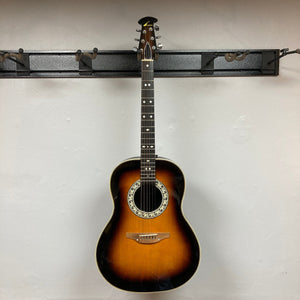 A 1976 Ovation 1112-1 Acoustic Guitar w/Case, made in the USA by Ovation, hangs on a wall-mounted rack against a plain white background.