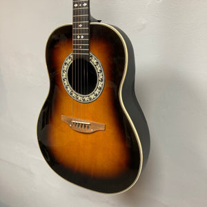 A 1976 Ovation 1112-1 USA-made acoustic guitar by Ovation, featuring six strings and decorative rosette, hangs on a white wall. Includes case.
