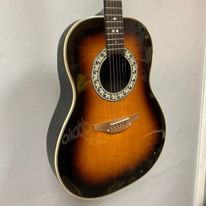 A 1976 Ovation 1112-1 Acoustic Guitar w/Case USA Consign hangs on a light-colored wall, featuring a sunburst finish, decorative rosette, and classic Ovation Lyrachord rounded back.