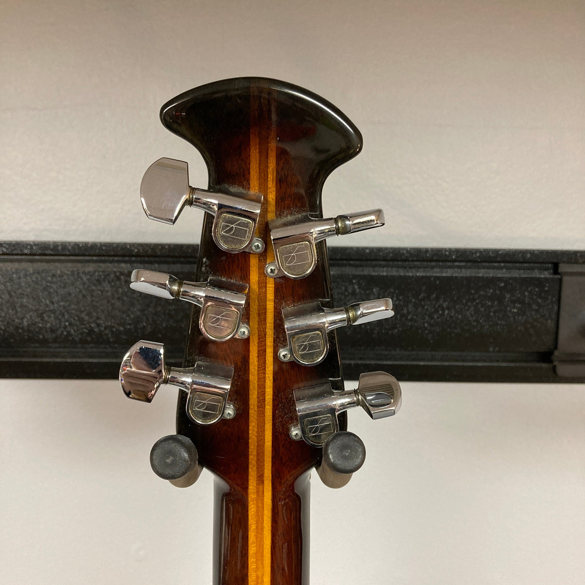 Close-up of the back of a 1976 Ovation 1112-1 Acoustic Guitar headstock, made in the USA, showing six silver tuners and brown wood with a central stripe. The Ovation rests on a black wall mount.