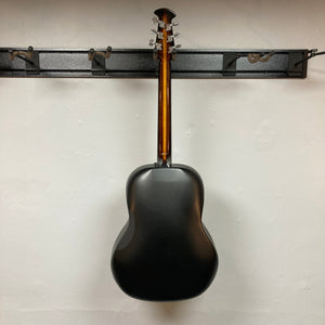 A 1976 Ovation 1112-1 Acoustic Guitar w/Case USA Consign is hanging by its headstock on a wall-mounted rack, displaying its curved Lyrachord back against a plain white wall.