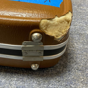 Close-up of a brown hard case for a 1976 Ovation 1112-1 Acoustic Guitar (USA), showing a damaged corner with yellow foam exposed; metal hinge and latches can be seen on blue-gray carpet.