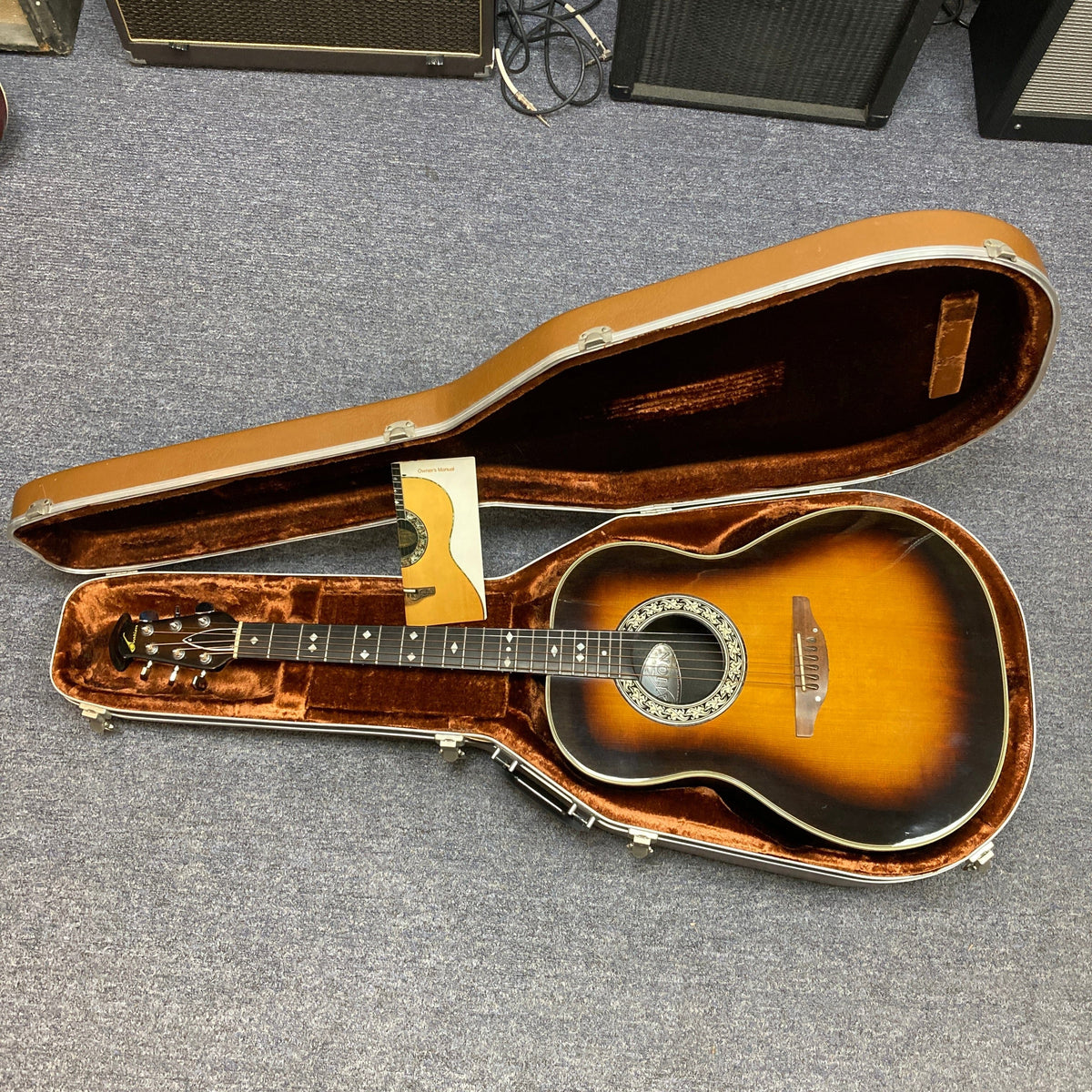 A 1976 Ovation 1112-1 acoustic guitar with decorative soundhole and Lyrachord rounded back is displayed in an open brown hard case on a gray carpet. An instructional booklet is included. Brand: Ovation.