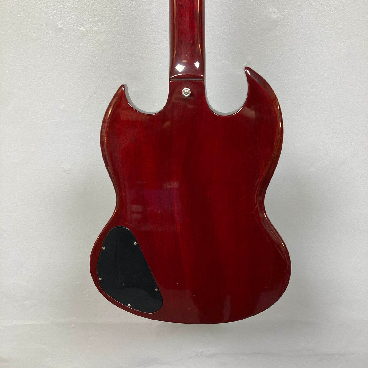 The back of a 1977 Gibson SG Standard Cherry electric guitar by Gibson is shown against a white wall, highlighting its double cutaway mahogany body. A black plastic control cover appears on the lower left side, echoing the classic style of the Gibson Kalamazoo era.