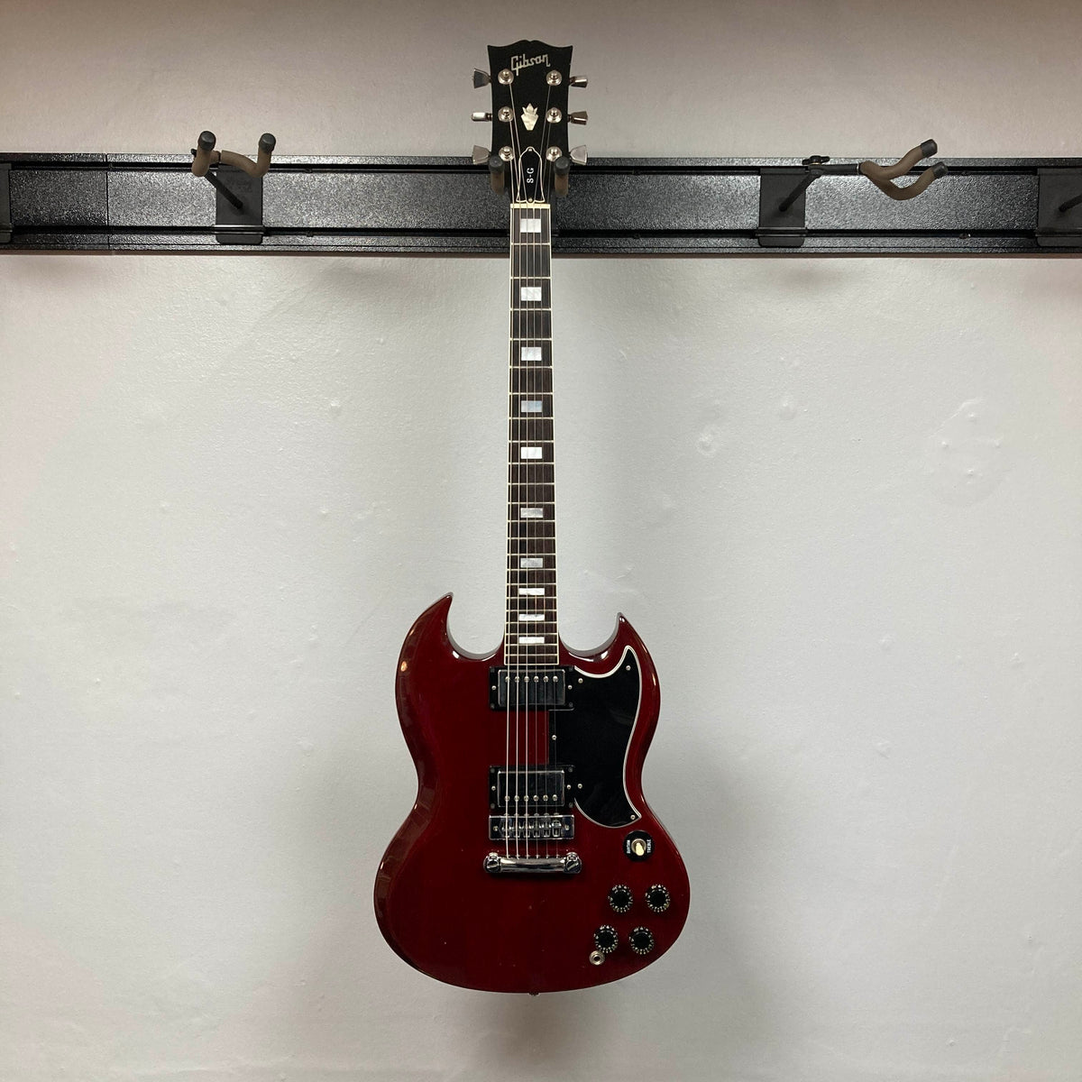 A 1977 Gibson SG Standard Cherry electric guitar by Gibson, featuring a mahogany body, black pickguard, and chrome hardware, hangs on a wall-mounted guitar holder against a white wall.