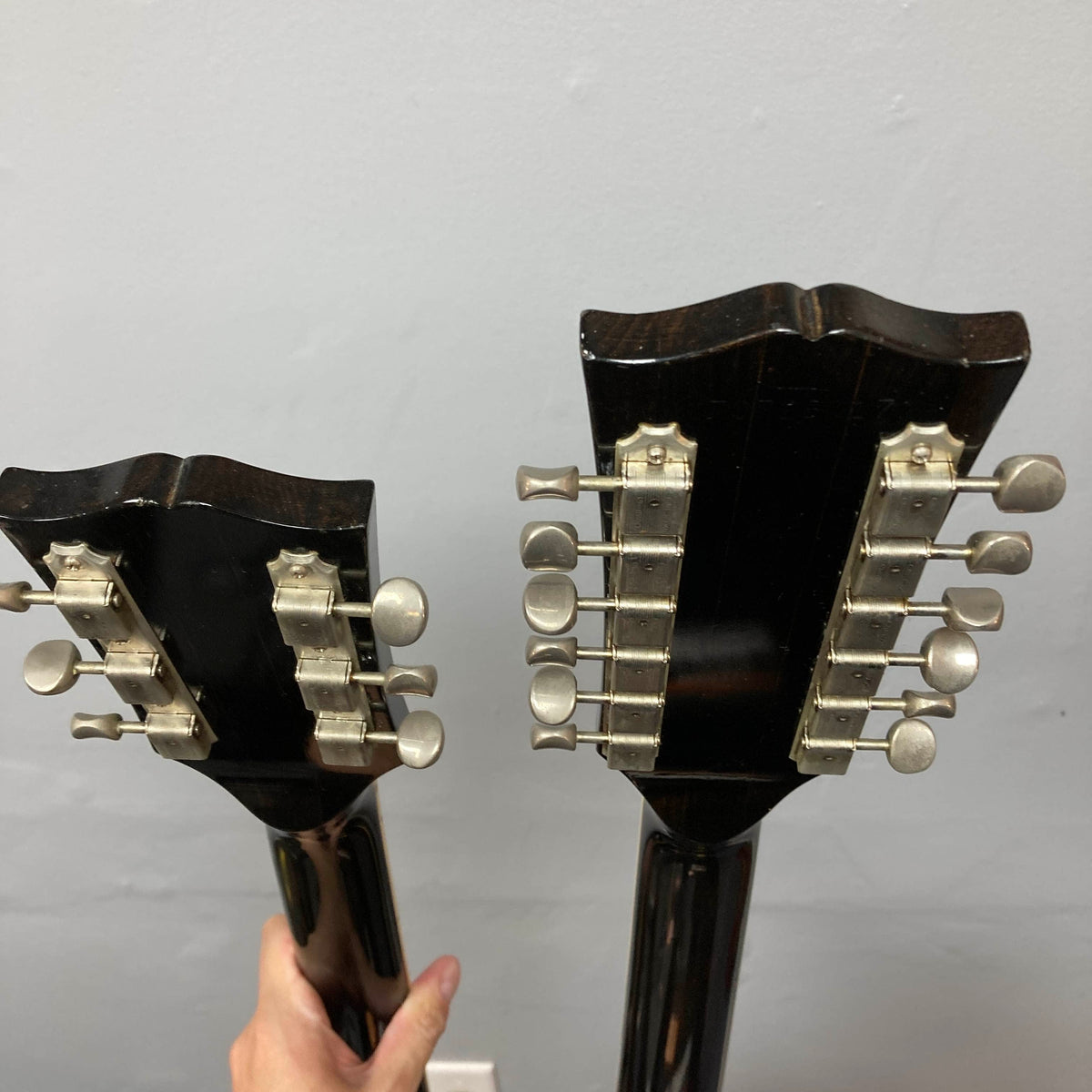 Two guitar headstocks with tuning pegs, one on the left and one on the right, are shown against a light gray wall. A hand holds the left guitar neck, which appears to be from a Gibson 1978 EDS-1275 Double Neck Walnut w/Case.