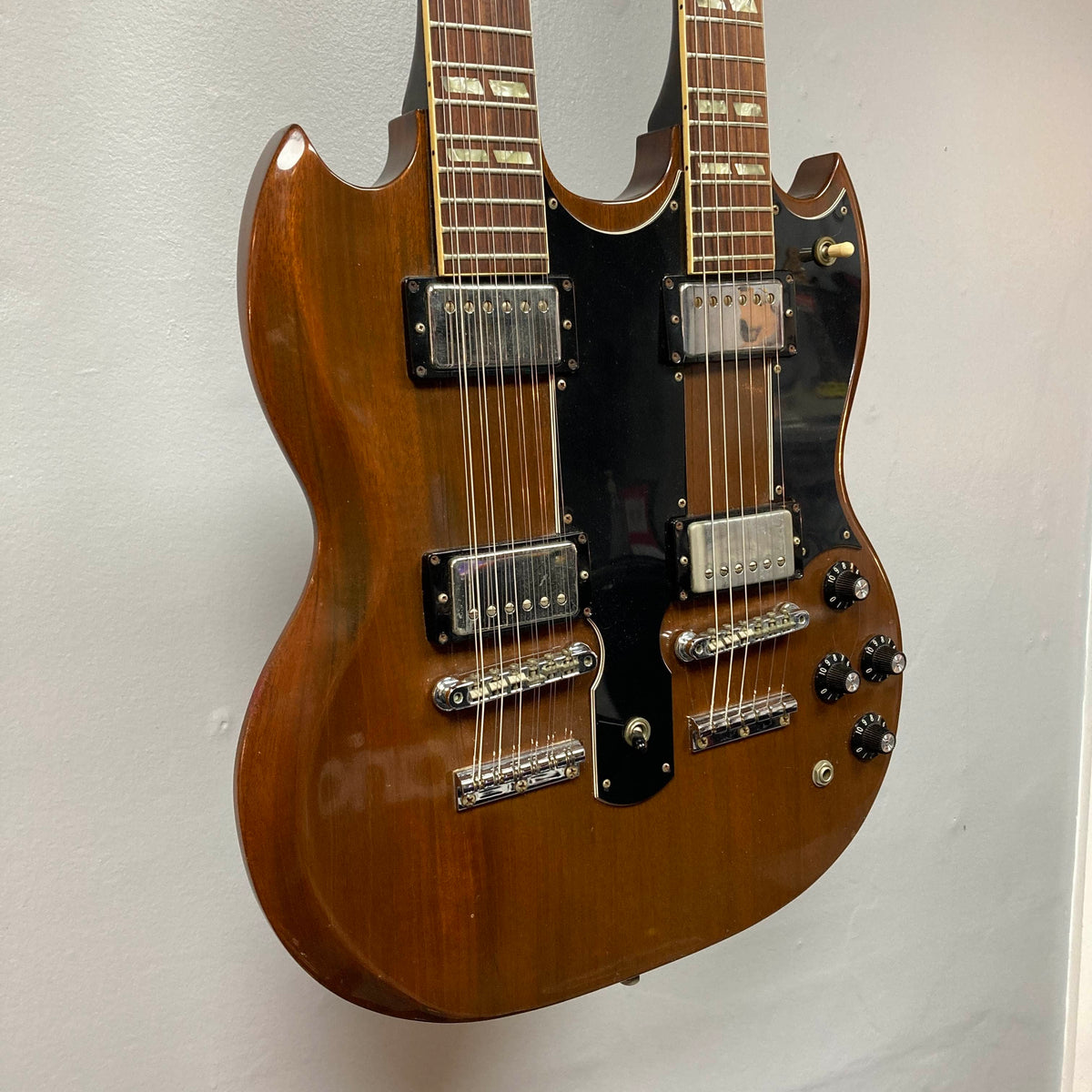 A 1978 Gibson EDS-1275 Double Neck electric guitar in a walnut finish, featuring a black pickguard, chrome hardware, and multiple control knobs, is displayed hanging on a white wall.