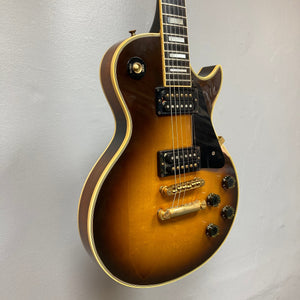 A close-up of a 1980 Gibson Les Paul Custom in Tobacco Sunburst, featuring gold hardware, black pickguard, and four control knobs, hangs against a plain light wall. Includes original hardshell case (OHSC).