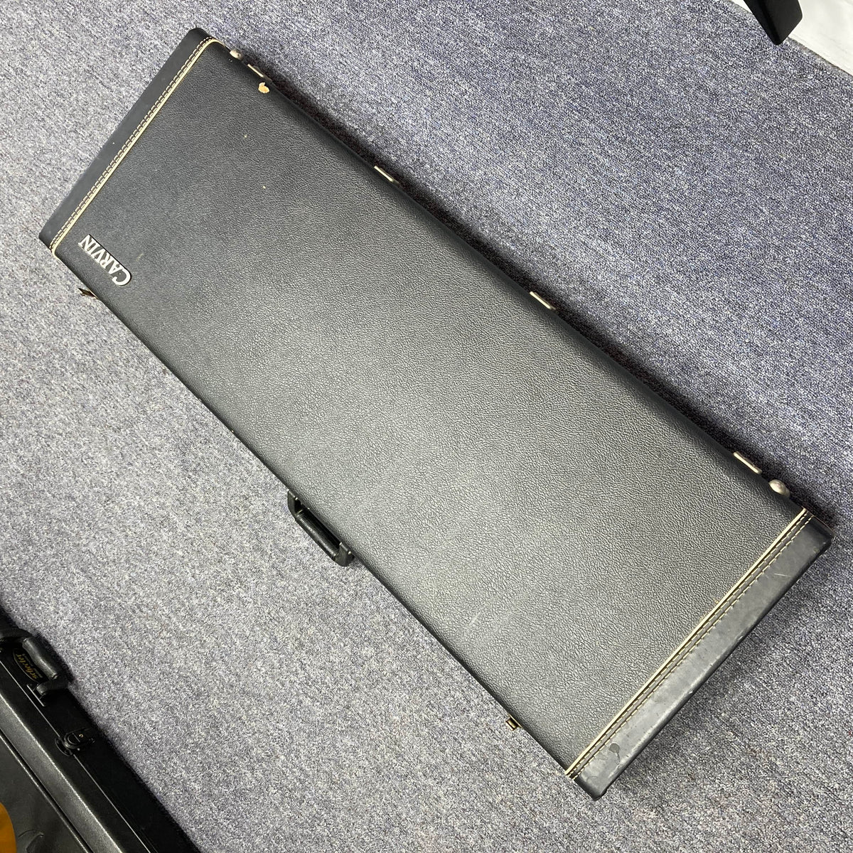 A closed black Carvin guitar case with white stitching, likely holding a 1992 Carvin Ultra V USA – Kahler Steeler Trem, MJ-Wound Seymour Duncan DCJ electric guitar, rests on a gray carpeted floor.