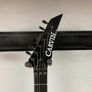 Headstock of a Carvin 1992 Ultra V USA with six tuners, black hardware, Kahler Steeler Trem, MJ‑wound Seymour Duncan DCJ pickup, and logo, shown against a white wall with a black horizontal bar.