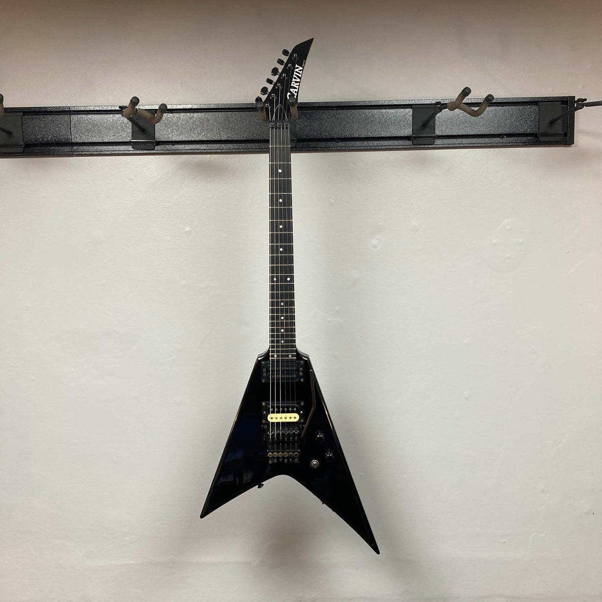 A black, USA-made 1992 Carvin Ultra V with a V-shaped body, Kahler Steeler tremolo, and MJ-wound Seymour Duncan DCJ humbucker hangs on a wall-mounted rack against a white wall.