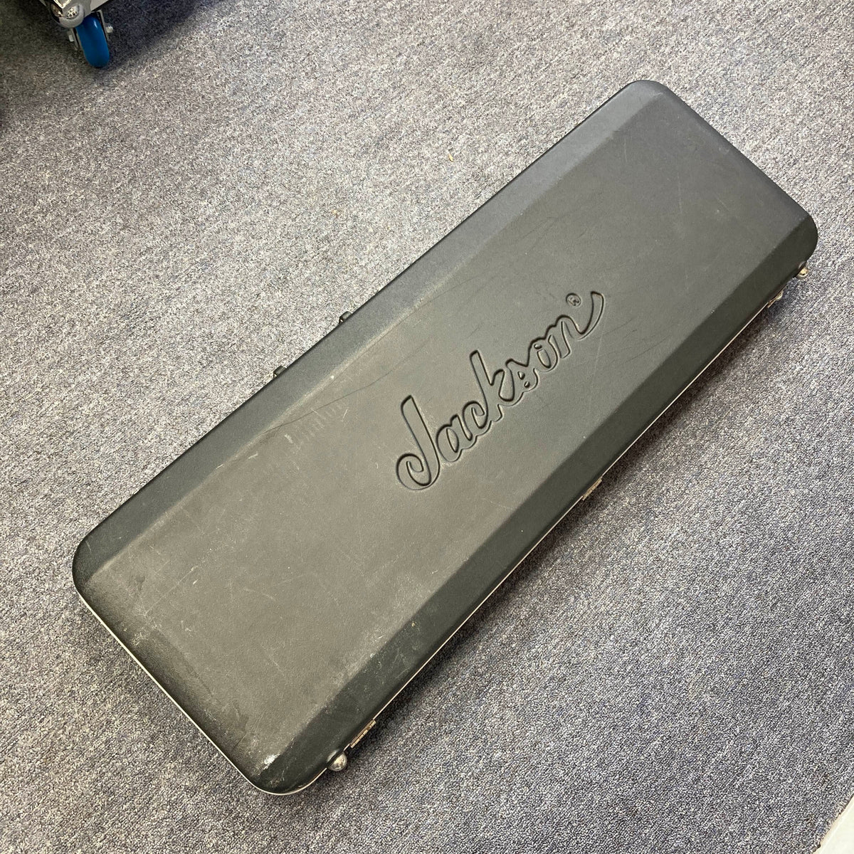 A black Jackson hard guitar case, featuring the “Jackson” logo and made for the 1995 Jackson SL1 Soloist Custom Shop Eerie Dess Swirl w/Case, is lying closed on a gray carpeted floor.