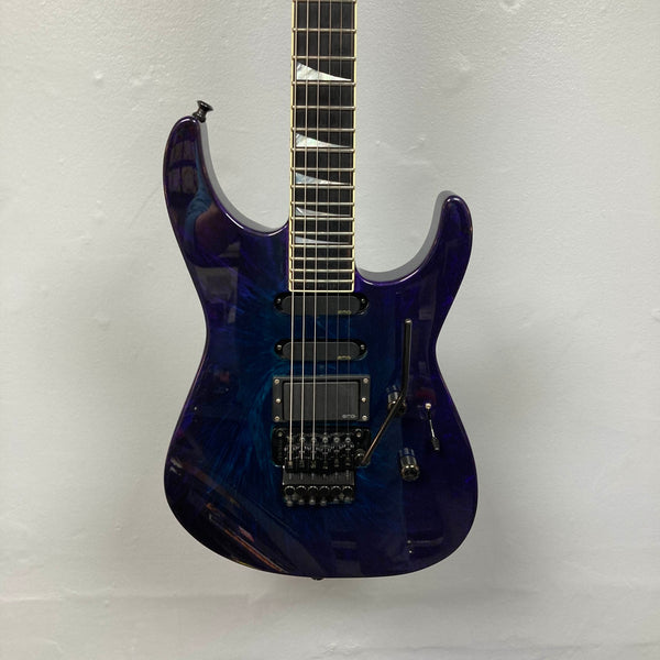 Hanging on the wall is a 1995 Jackson SL1 Soloist Custom Shop Eerie Dess Swirl electric guitar, featuring a striking purple and blue burst finish, black pickups, a Floyd Rose tremolo bridge, and a sleek maple neck—an iconic model from Jackson’s Custom Shop collection.