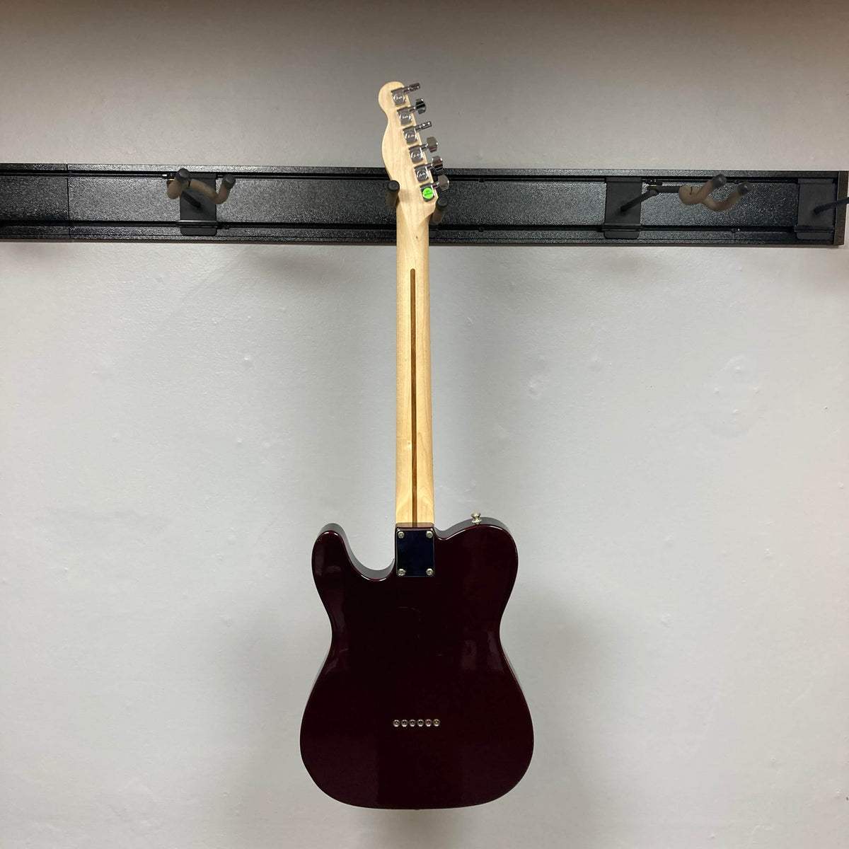 A 2007 Fender Standard Telecaster MIM in Midnight Wine Red by Fender hangs on a black wall rack, shown from the back. The light-colored neck and tuning pegs are visible; pickups and front details are not. The wall is plain and off-white.