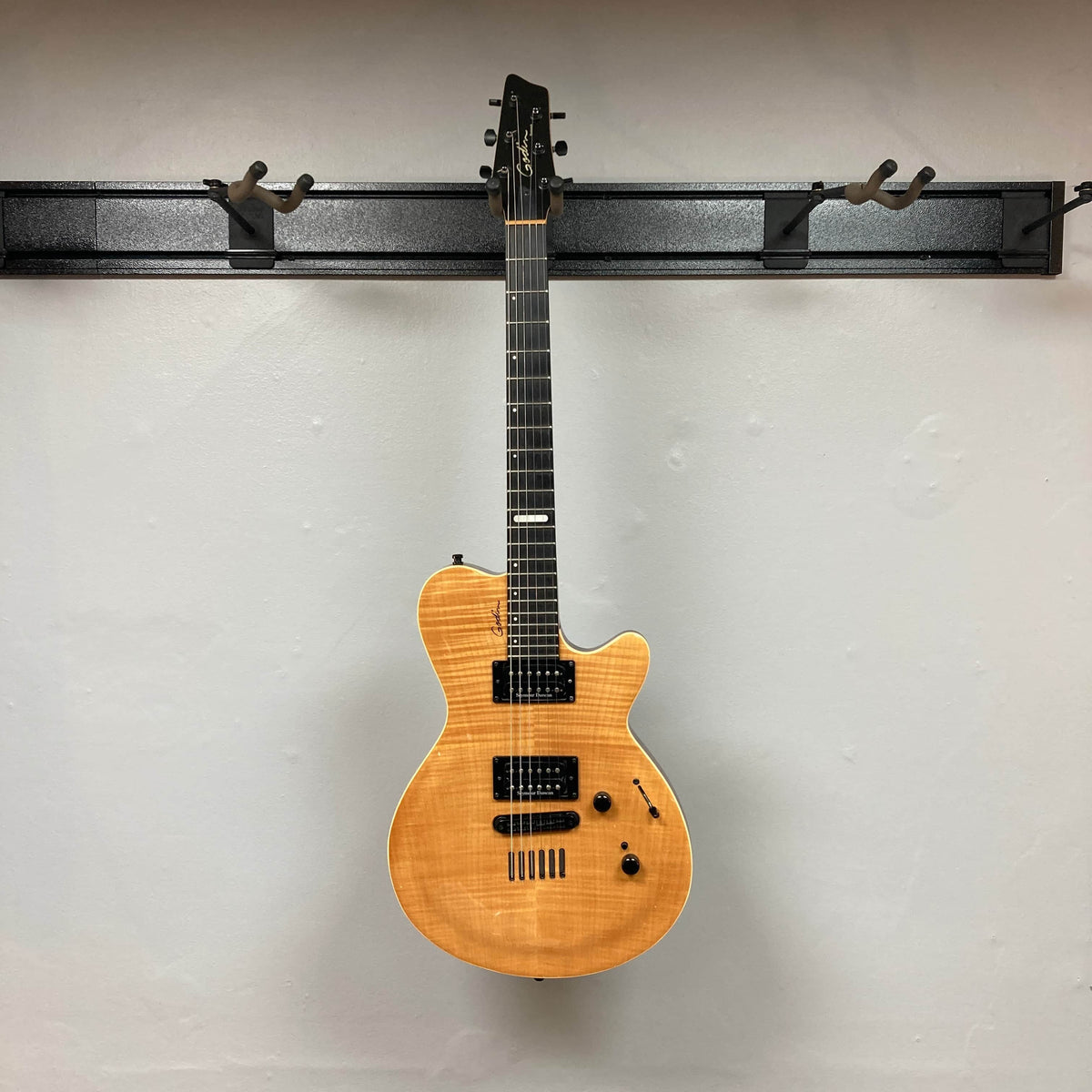 A 2009 Godin Summit CT with a carved flame maple cap by Godin, featuring a natural wood finish, hangs on a wall-mounted rack with several empty hooks.