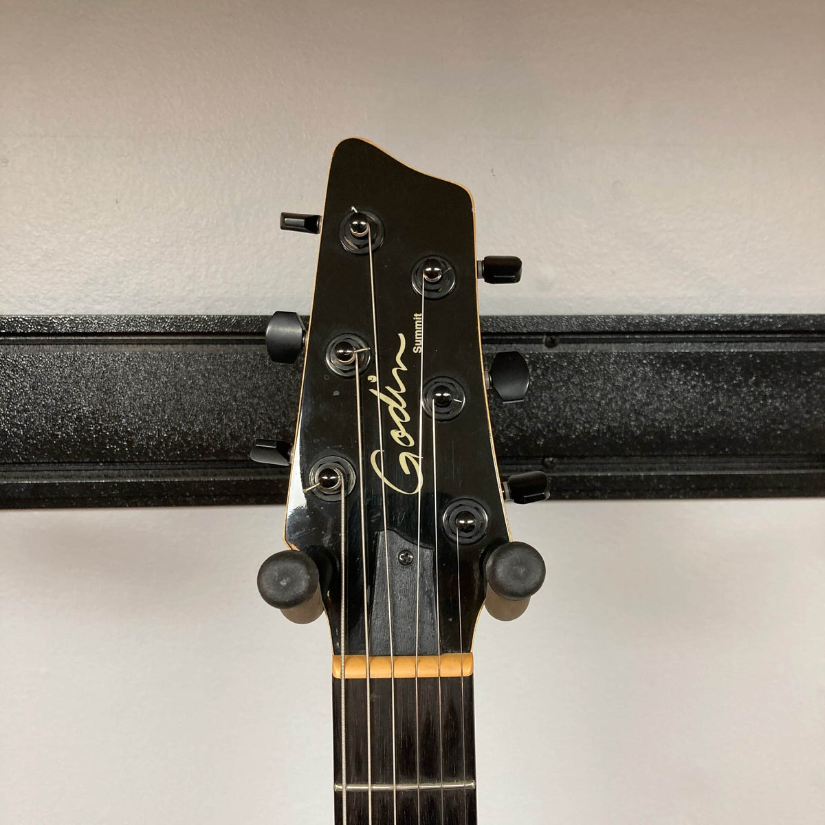 Headstock of a 2009 Godin Summit CT with six tuning pegs, mounted on a black wall hanger against a light wall—an ideal display piece for fans of flame maple Godin electric guitars.