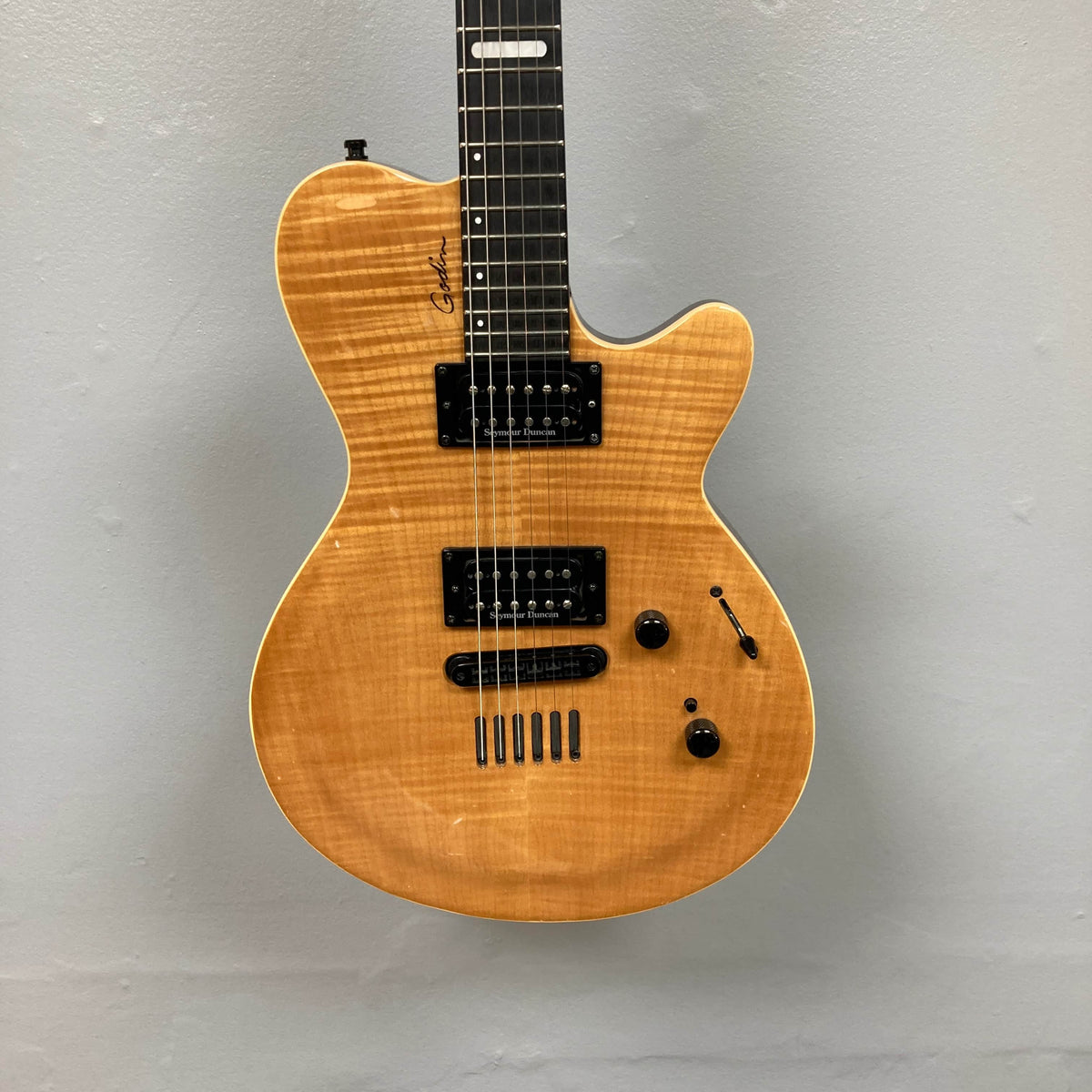 A 2009 Godin Summit CT electric guitar with a carved flame maple cap, Seymour Duncan humbuckers, three knobs, and a toggle switch hangs against a plain gray wall.