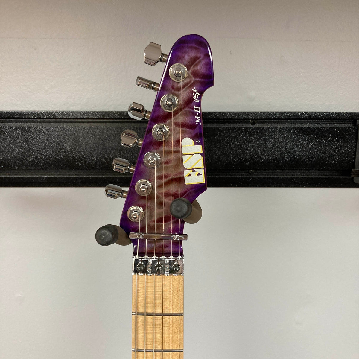 A close-up of the headstock of a 2019 ESP USA M-II Purple Haze Burst electric guitar reveals six tuning pegs, string guides, and part of the wooden fretboard, all showcased in a striking Purple Haze Burst finish against a white wall. This American-made ESP guitar exemplifies exceptional craftsmanship and an eye-catching design, and comes complete with its original case.