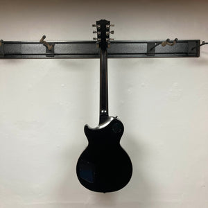 The 2025 Gibson Les Paul Studio Modern in Smokehouse Satin by Gibson hangs by its neck on a wall-mounted rack, viewed from the back against a plain white wall, with its Gator Molded Case not shown.