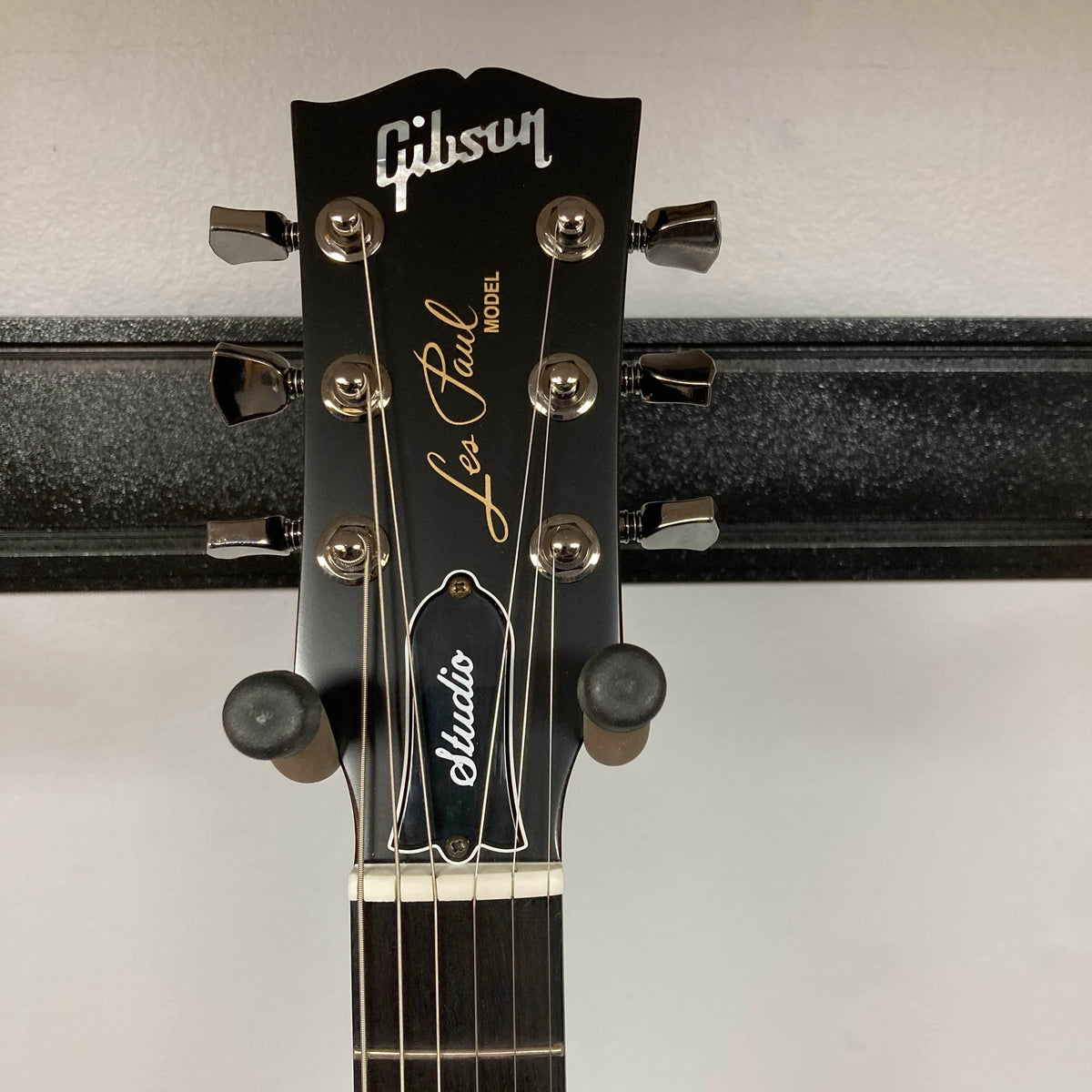 Close-up of the headstock of the 2025 Gibson Les Paul Studio Modern in Smokehouse Satin, showing tuning pegs, logo, and model name against a wall-mounted guitar hanger. Includes Gator Molded Case.
