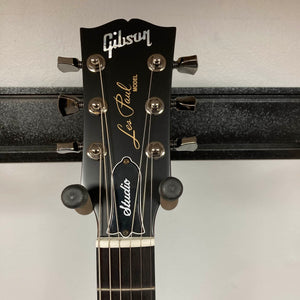 Close-up of the headstock of the 2025 Gibson Les Paul Studio Modern in Smokehouse Satin, showing tuning pegs, logo, and model name against a wall-mounted guitar hanger. Includes Gator Molded Case.
