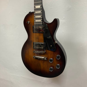 The 2025 Gibson Les Paul Studio Modern in Smokehouse Satin, featuring dual humbuckers and four control knobs, hangs elegantly on a white wall; includes a Gator Molded Case.