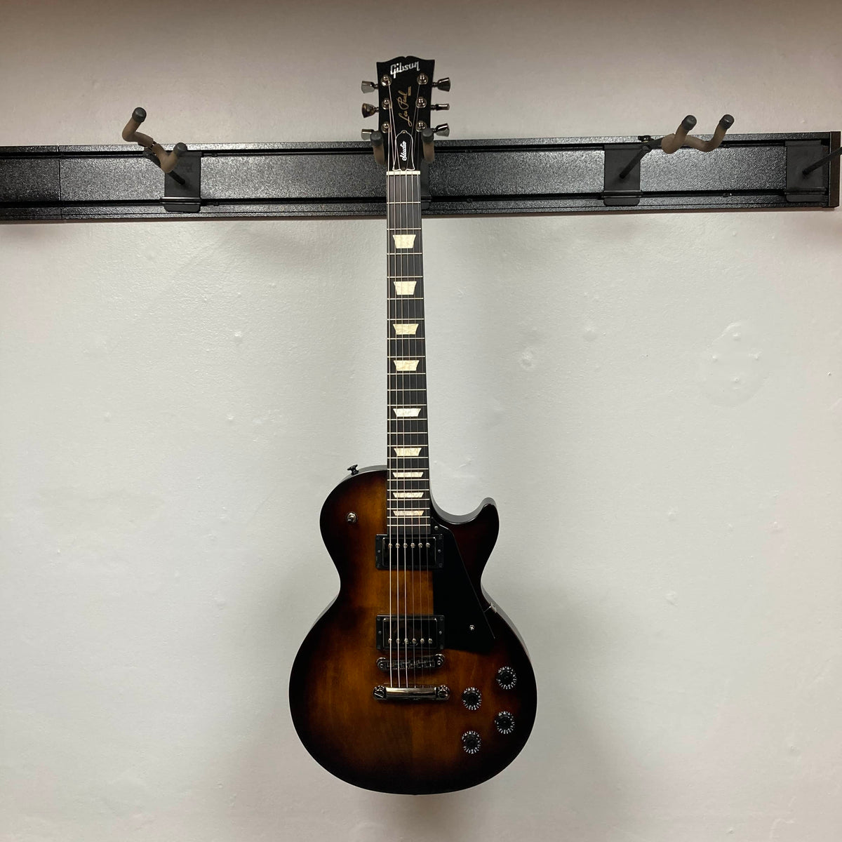 A 2025 Gibson Les Paul Studio Modern in Smokehouse Satin hangs on a wall-mounted rack against a white background, highlighting its sleek single cutaway design and comes with a Gator Molded Case.