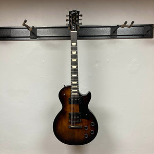 A 2025 Gibson Les Paul Studio Modern in Smokehouse Satin hangs on a wall-mounted rack against a white background, highlighting its sleek single cutaway design and comes with a Gator Molded Case.