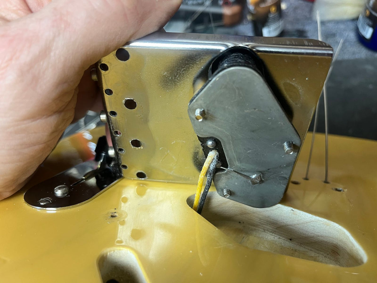 A hand holds a Fender Custom Shop '51 Reissue Nocaster NOS 2006 metal bridge with pickup above a yellow Fender electric guitar body during installation or repair.