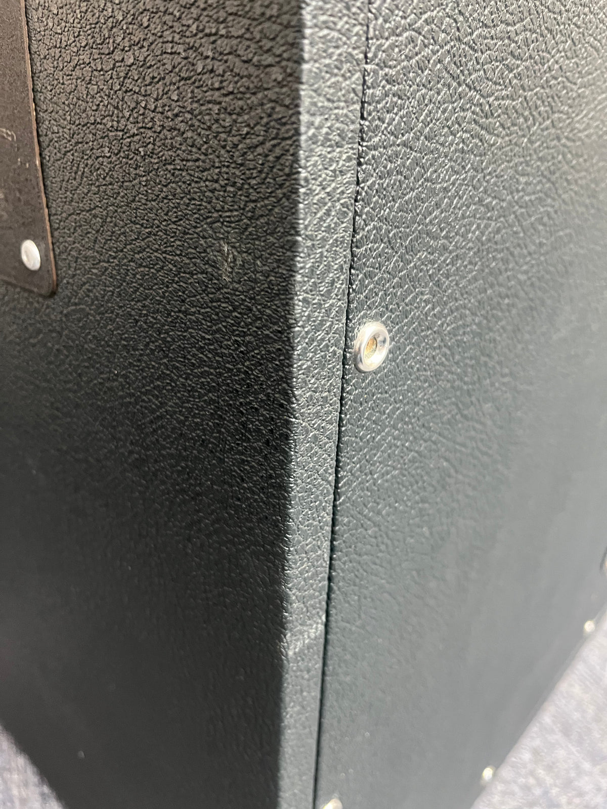Close-up of a black textured box or case featuring visible metal screws on the corners and side panels, reminiscent of the rugged design found on a vintage 1969 Marshall 4x12 Cabinet with Matching Celestion T1281 Greenbacks and classic basketweave grille cloth.