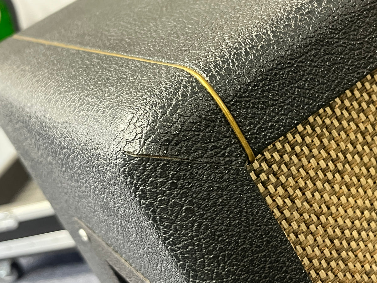 Close-up of a corner of a textured black 1969 Marshall 4x12 Cabinet with matching Celestion T1281 Greenbacks, showing a small tear in the covering and visible gold piping; partially visible woven basketweave grille cloth.