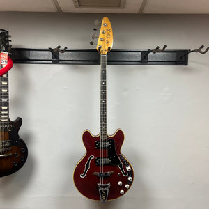 A red 1967-68 Vox Sidewinder IV Bass (V272) Cherry is hanging on a wall-mounted rack, its natural finish headstock visible next to a partially shown vintage Italian bass.