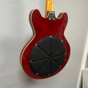 A cherry red 1967-68 Vox Sidewinder IV Bass (V272) by Vox hangs on the wall, its back featuring a round, padded black cover secured with visible screws—a detail echoing classic Italian bass design.