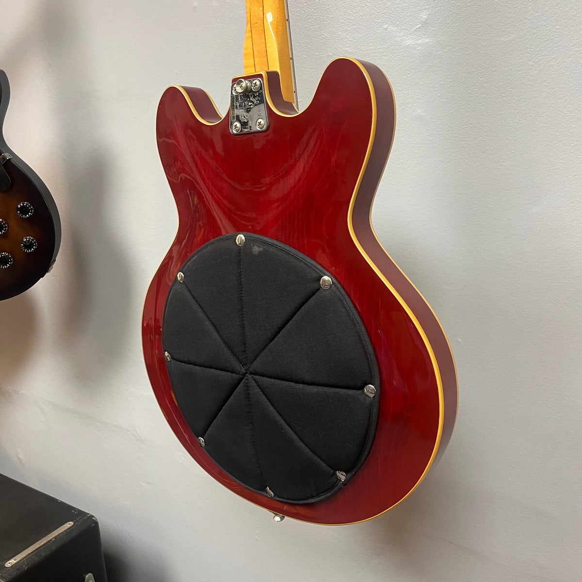 A 1967-68 Vox Sidewinder IV Bass (V272) in cherry finish hangs on the wall, showcasing a large black circular pad with visible screws on the back—classic of vintage Italian Vox bass designs.