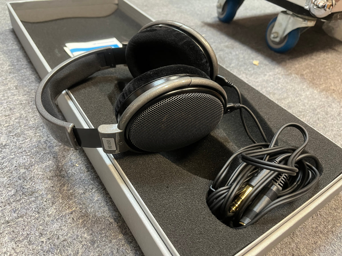 A pair of preowned Sennheiser HD 650 Reference Class headphones, featuring an open-back black design with a coiled cable and adapter, are displayed in a foam-lined case on a grey carpeted surface.