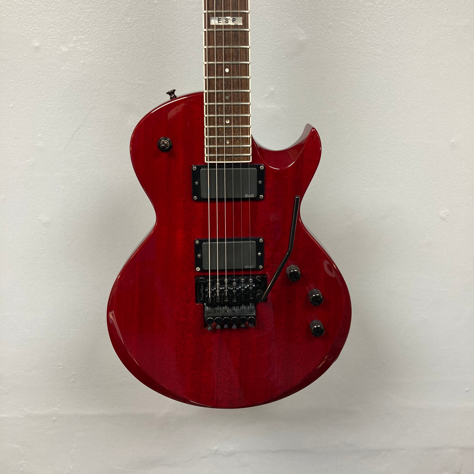 An ESP Standard Eclipse Transparent Cherry Red electric guitar with dual humbuckers, tremolo bridge, and four knobs is shown against a plain white wall. Includes case. Brand: Esp. An ESP Standard Eclipse Transparent Cherry Red electric guitar with dual humbuckers, tremolo bridge, and four knobs is shown against a plain white wall. Includes case. Brand: Esp.