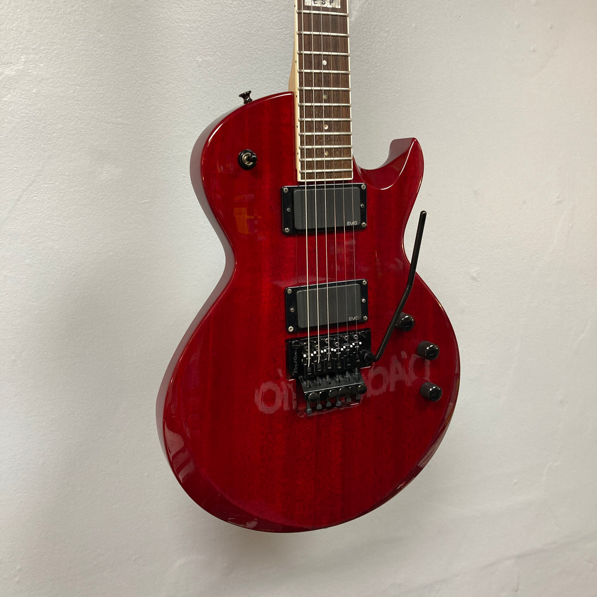 An ESP Standard Eclipse Transparent Cherry Red electric guitar by Esp, with case, dual humbuckers, and tremolo bridge, hangs against a plain white wall.