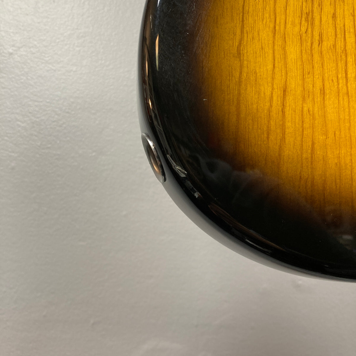 Close-up of the side of a Fender Sting P Bass Sunburst electric bass by Fender, highlighting the strap button with a plain light-colored wall in the background.
