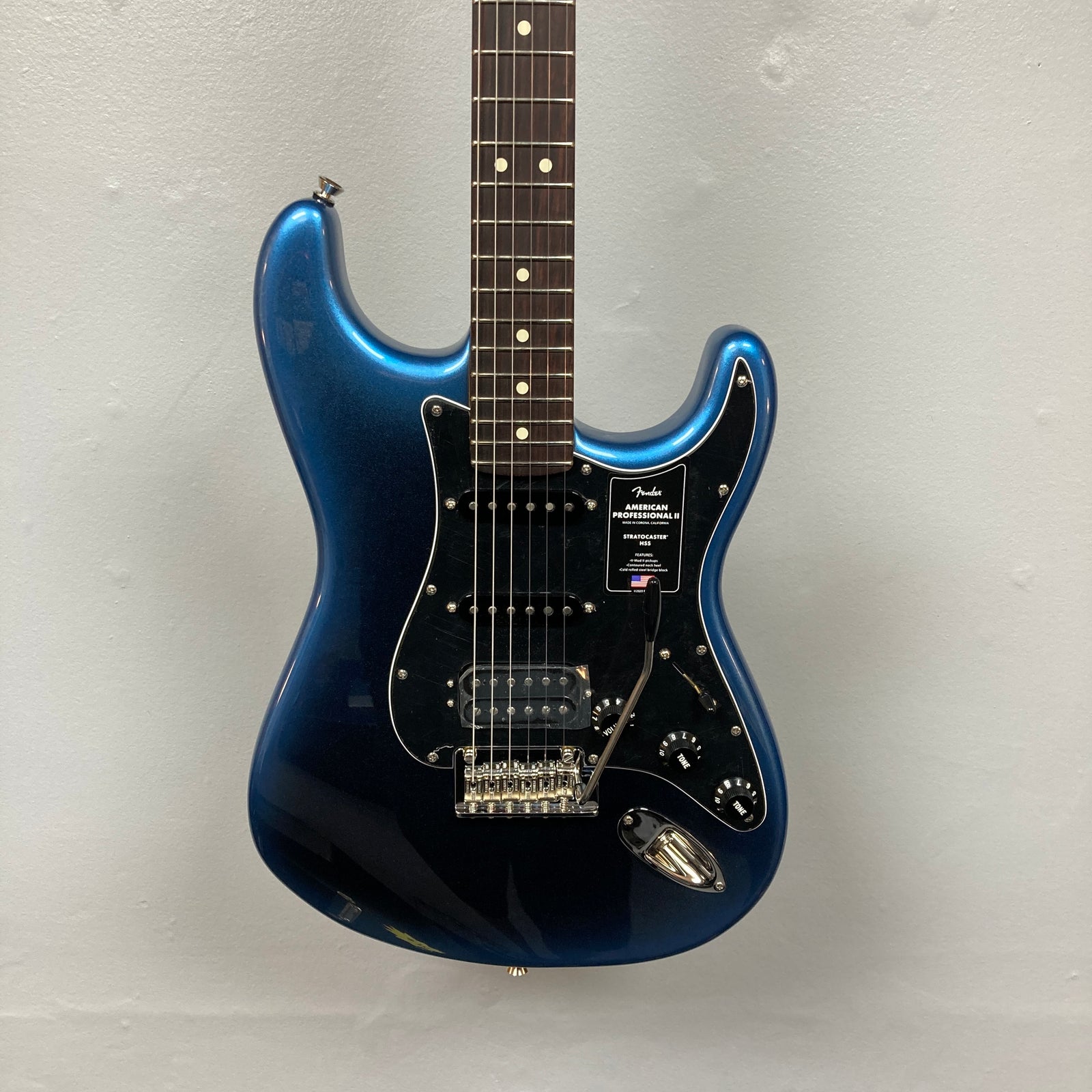 A Fender American Professional II Stratocaster HSS in Dark Night with case, featuring a black pickguard, photographed against a plain light gray wall. A Fender American Professional II Stratocaster HSS in Dark Night with case, featuring a black pickguard, photographed against a plain light gray wall.